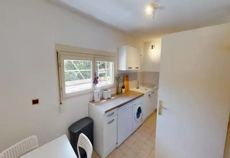 Cuisine compacte et rangée avec plan de travail en bois, électroménagers sous-plan (lave-linge/lave-vaisselle), évier et rangements muraux. La lumière naturelle entre par la fenêtre au-dessus du comptoir, créant un espace fonctionnel et lumineux.