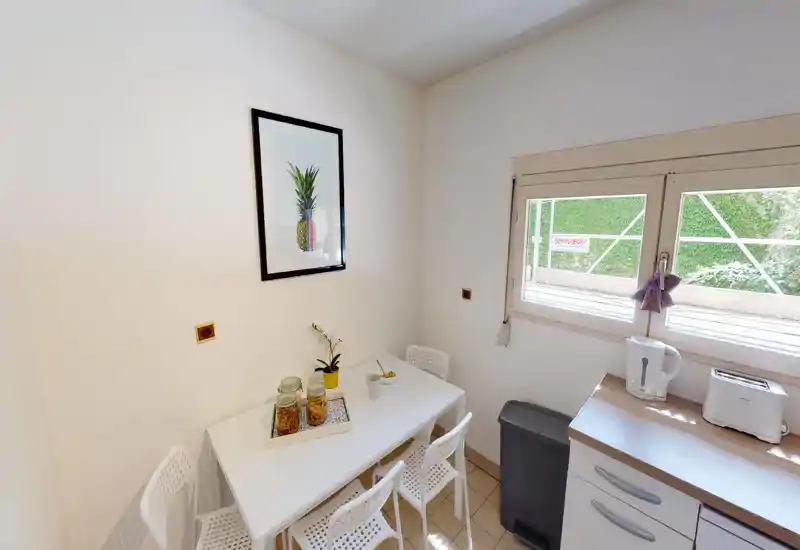 Coin cuisine/salle à manger lumineux avec une table blanche pour quatre, des appareils sur le plan de travail (bouilloire et grille-pain) et une fenêtre laissant entrer la lumière naturelle. Décor épuré et minimaliste avec un cadre et de petites plantes décoratives.