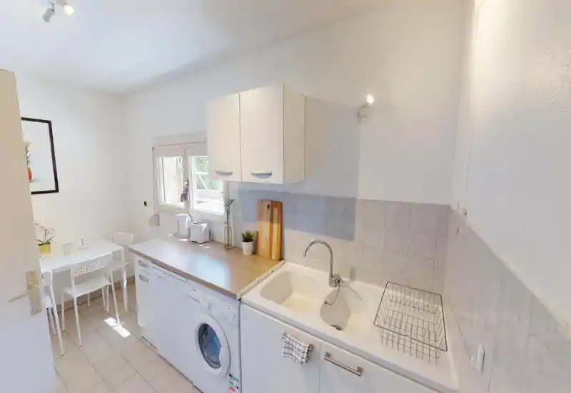 Compact, bright kitchen with a double sink, wooden countertop, washing machine and a small dining nook by the window — neat, functional and well-lit.