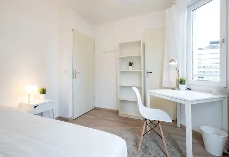 Bright, minimal bedroom with a visible bed, bedside table and a desk by the window. The room is clean and simply furnished with natural light and small plants for a welcoming feel.