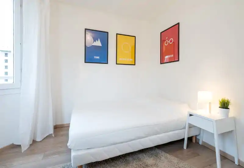 Bright, minimalist bedroom with a white double bed, bedside table with lamp and plant, and three colorful framed prints above the bed—ideal for a clean, modern rental listing.