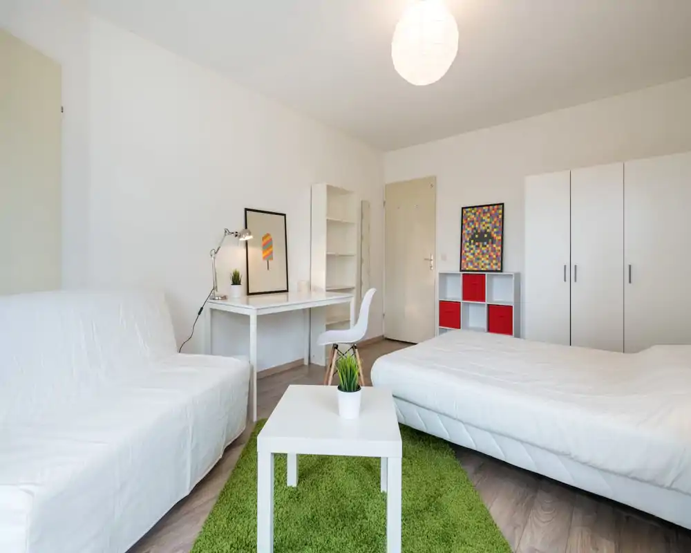 Camera da letto luminosa e moderna con un ampio letto bianco, divano, scrivania e mobili contenitori. Arredo minimal con tappeto verde e tocchi di colore per un ambiente fresco e accogliente.