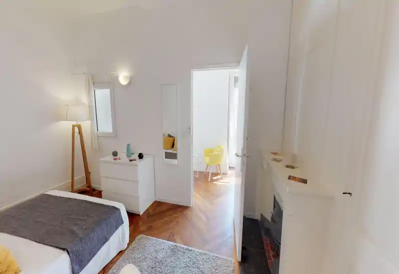 Bright, softly styled bedroom with a large bed, wooden herringbone floor and warm accents. The room is well-lit with natural light and tasteful textiles for a cozy feel.