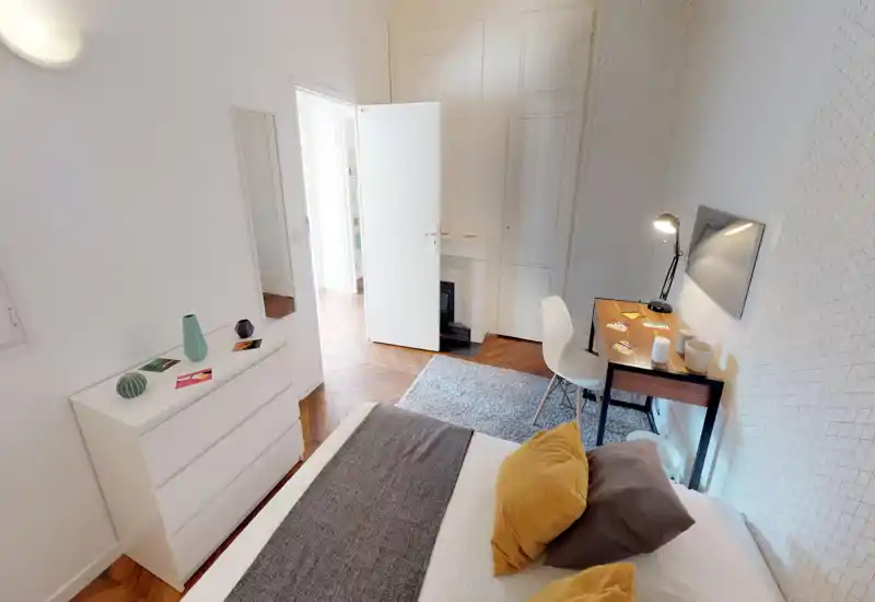 Bright, well-staged bedroom with a comfortable bed in the foreground, decorative pillows and a small working desk by the wall. White walls, parquet floor and a soft rug create a warm, inviting atmosphere ideal for rental photos.