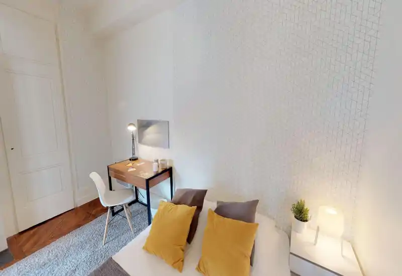 Bright, modern bedroom with a neatly made bed, warm mustard cushions, a small bedside table with lamp and a compact wooden desk with chair — calm and well-staged for guests.