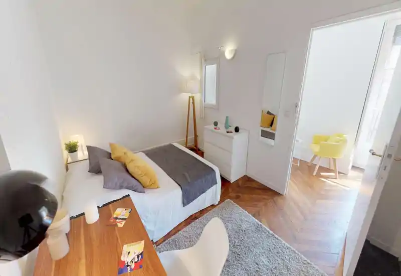 Bright, stylish bedroom with a double bed, layered textiles and warm wooden floors; natural light enters from the adjacent room creating an inviting atmosphere.