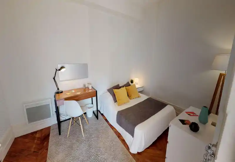 Bright, neatly styled bedroom with a double bed dressed in white linen, decorative throw and cushions, plus a small desk and bedside lighting — ideal for comfortable stays.