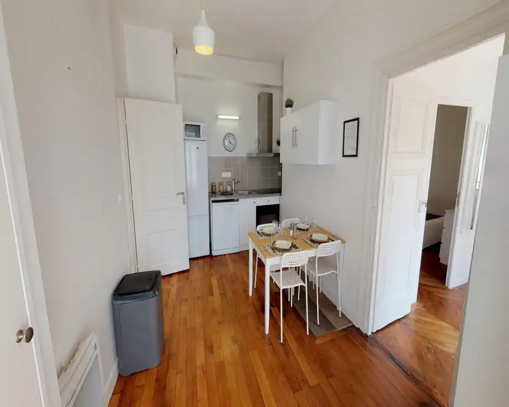 Cocina abierta y compacta con electrodomésticos integrados y una mesa pequeña para cuatro. Espacio luminoso y ordenado con suelos de madera y muebles blancos modernos, ideal para un piso urbano.