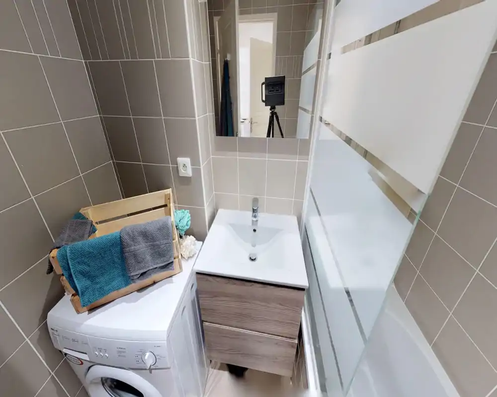 Baño moderno y compacto con lavabo, mueble, espejo, bañera con mampara de vidrio esmerilado y lavadora colocada de forma ordenada para optimizar el espacio.
