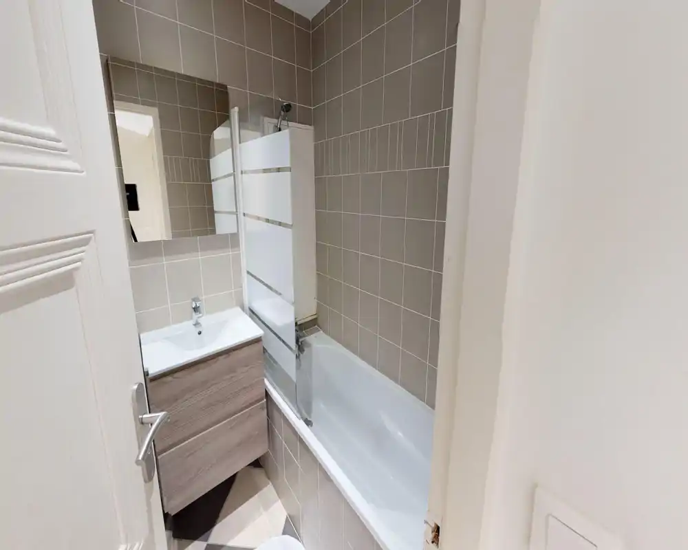 Baño pequeño moderno con bañera y mueble lavabo; azulejos grises neutros y mampara de cristal esmerilado que aportan un aspecto limpio y contemporáneo.