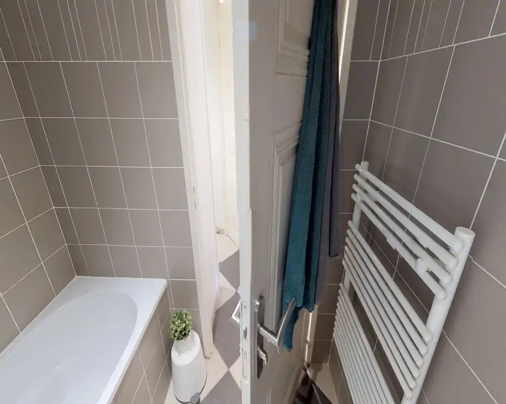 Baño moderno y compacto con paredes alicatadas, bañera, toallero calefactado y decoración neutra — luminoso y limpio.