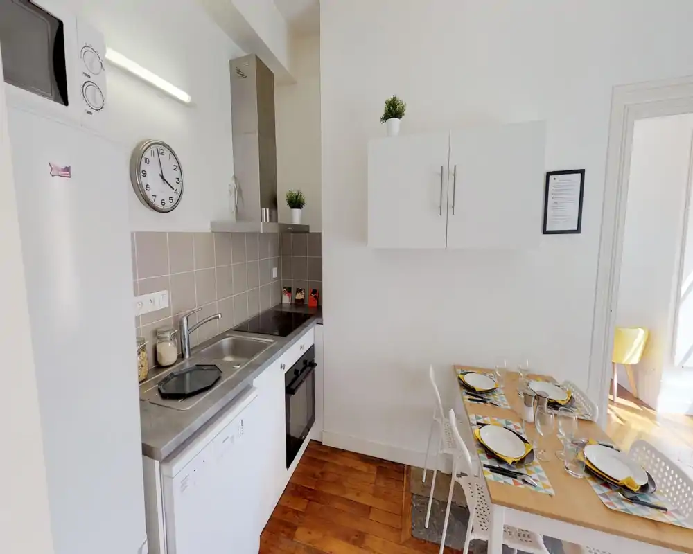 Cocina compacta y bien equipada con fregadero de acero, placa y horno eléctricos, frigorífico y mesa para cuatro puesta. Estilo luminoso y limpio con pequeñas plantas y vajilla coordinada.