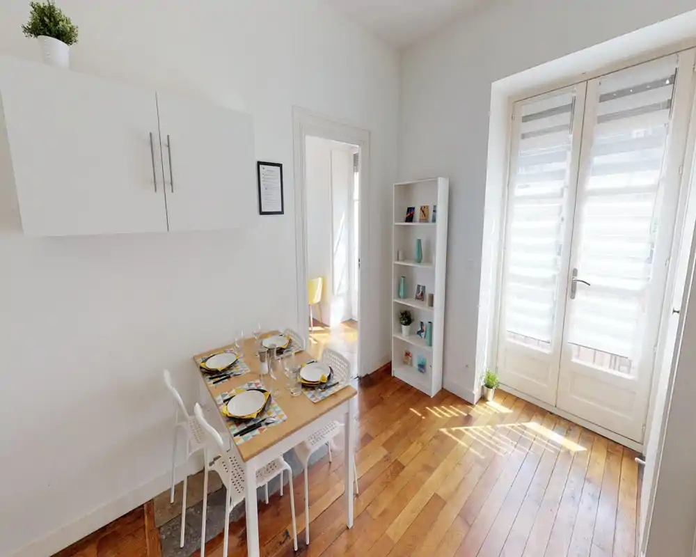 Zona de comedor luminosa y sencilla con mesa puesta para cuatro, suelos de madera y grandes puertas acristaladas que dejan entrar abundante luz natural.