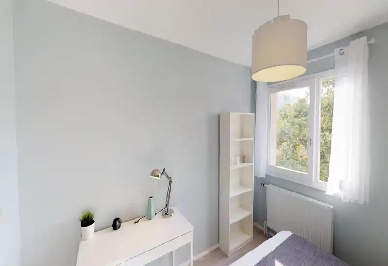 Camera da letto luminosa e minimale con pareti pastello chiare, una piccola scrivania e una libreria accanto a una grande finestra che lascia entrare luce naturale. L’inquadratura mostra l’arredo sobrio e la disposizione funzionale, ideale per soggiorni brevi.