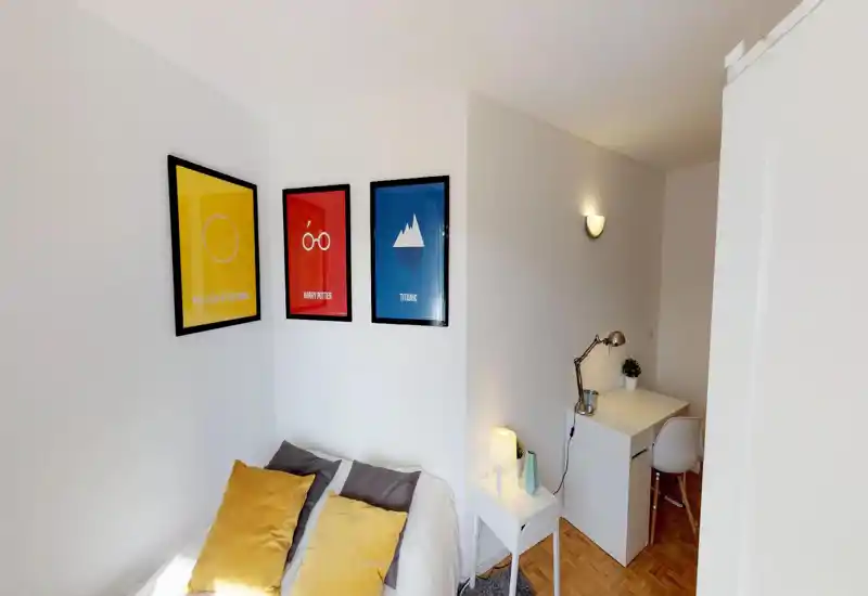 Small, bright bedroom with a neatly made bed, colorful cushions and three framed posters on the wall; a bedside table and a compact desk with lamp create a practical, cozy corner.