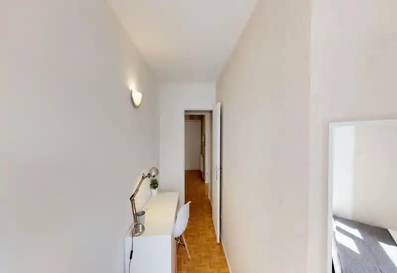 Narrow bedroom view with a workspace: a small desk, chair and lamp along the wall and a partial view of the bed at the right edge. Bright, minimal and neatly arranged, suitable for a single occupant or short-term stays.