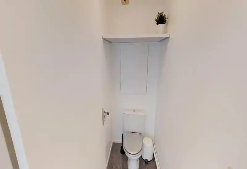 Petit WC privé avec toilette standard, petite poubelle et étagère avec plante ; murs blancs propres et sol neutre.