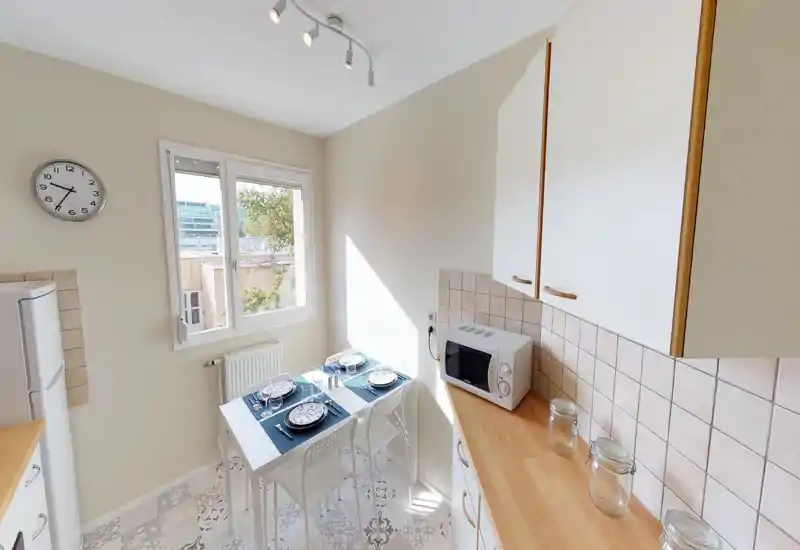 Petite cuisine lumineuse avec table dressée pour quatre, placards blancs, plans de travail en bois et micro-ondes — parfaite pour un appartement compact.