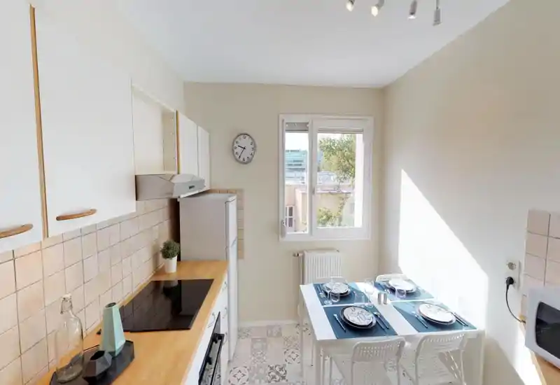 Cuisine compacte et lumineuse avec une petite table dressée pour quatre, meubles blancs, plan de travail en bois et carrelage à motifs. La lumière naturelle de la fenêtre apporte une atmosphère propre et accueillante.