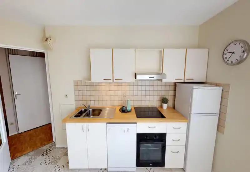 Cucina compatta con piano in legno, mobili bianchi, forno e lavastoviglie incassati, piano a induzione e frigorifero a colonna. Piastrelle neutre e arredo essenziale rendono lo spazio pulito e funzionale, ideale per un piccolo appartamento.