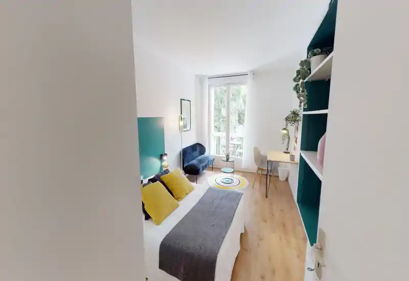Camera da letto luminosa e di stile con letto matrimoniale vestito in bianco, cuscini gialli come accento e un copriletto grigio. La stanza include un divanetto blu, una scrivania vicino alla finestra, scaffali decorati con piante e una grande finestra che illumina l’ambiente.