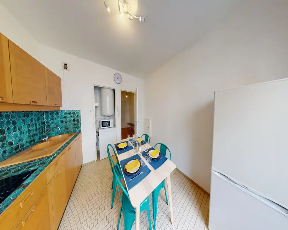 Bright furnished kitchen with a turquoise tile backsplash, wooden cabinets, small dining table set for four and a freestanding fridge — a clean, colorful space ideal for casual meals.