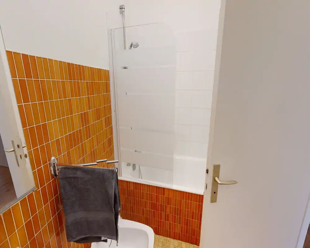 Compact bathroom with a bathtub and partial frosted glass shower screen, warm orange tiles and a towel rail; bright, well-lit space suitable for a rental listing.