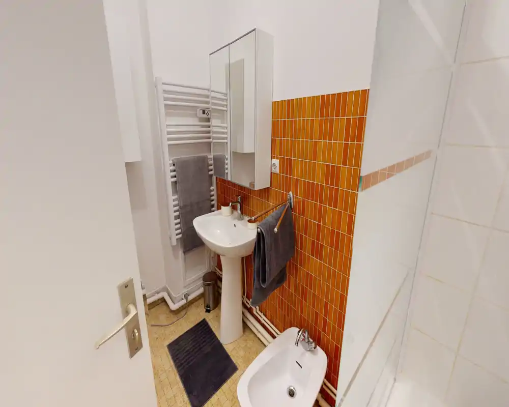 Compact, well-lit bathroom with a pedestal sink, bidet, mirrored cabinet and orange tiled accent wall. Clean towels and tidy fixtures make it practical for guests.