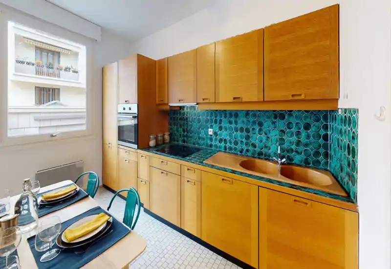Cucina luminosa in stile mid-century con pensili in legno caldo e un forte paraschizzi a piastrelle turchesi circolari, forno integrato, piano a induzione e lavello doppio; tavolo da pranzo per due vicino alla finestra.