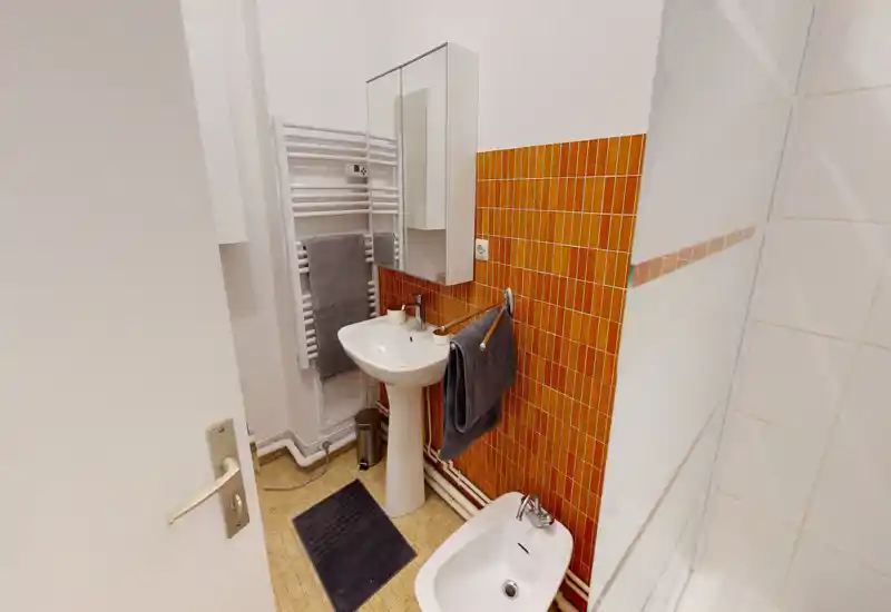 Compact, well-lit bathroom with a pedestal sink, bidet, mirrored cabinet and orange tiled accent wall. Clean towels and tidy fixtures make it practical for guests.