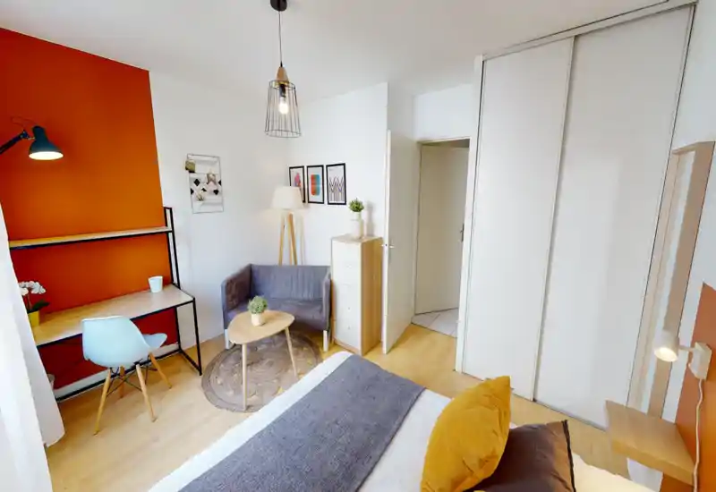 Bright, well-staged bedroom with a comfortable double bed, warm orange accent wall, desk area and small seating nook — ideal for a cozy rental stay.