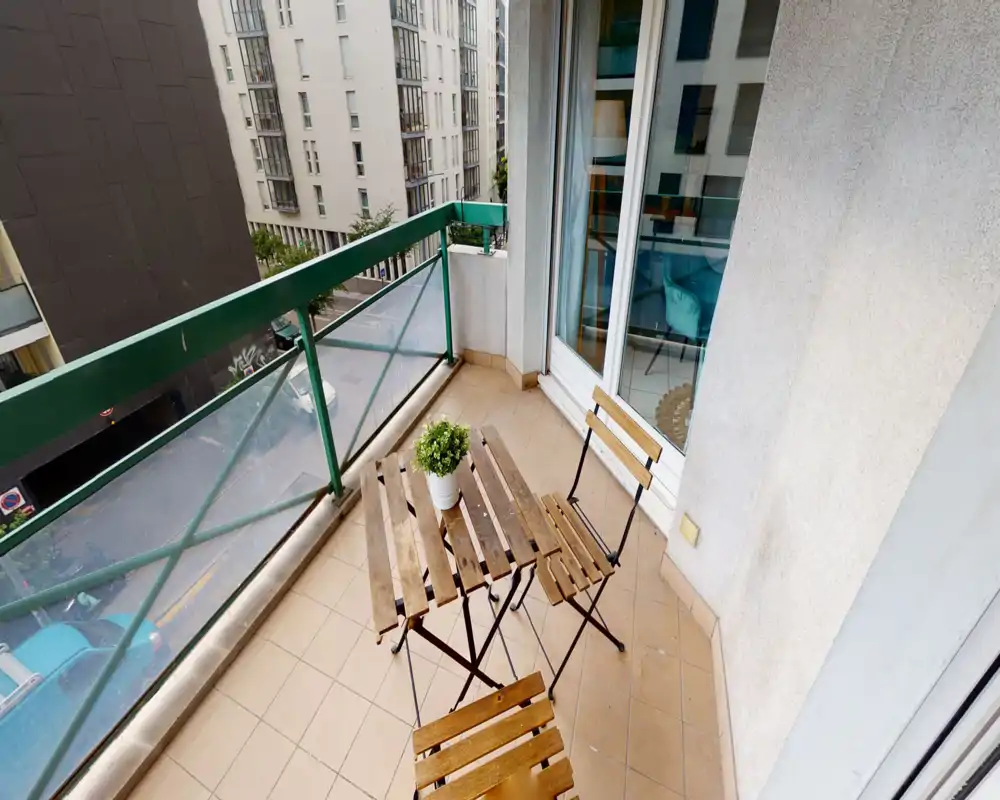 Sunny small balcony with a wooden bistro set and potted plant, glass-and-metal railing and tiled floor — perfect for morning coffee overlooking the street.