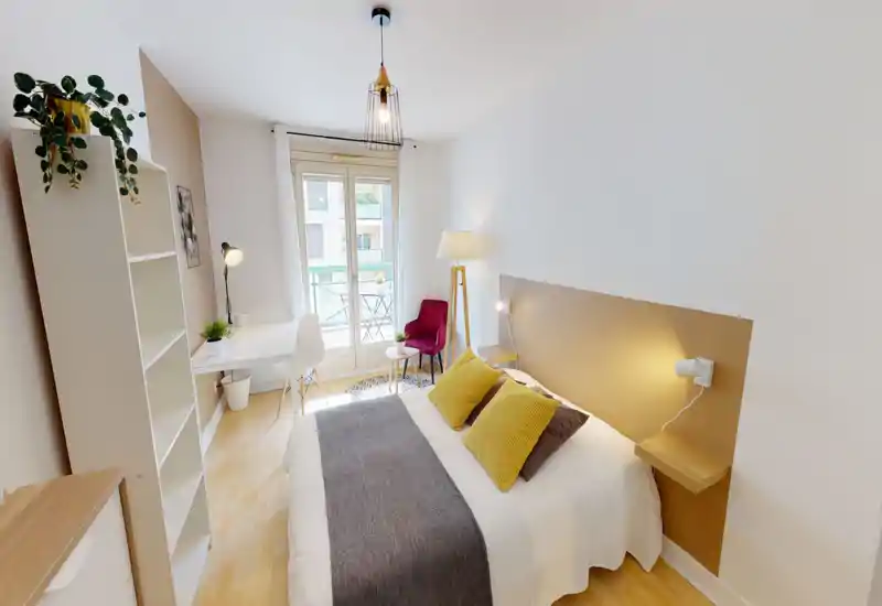 Chambre lumineuse et élégante avec un lit double aux coussins moutarde, un bureau près de la fenêtre et un coin salon donnant sur le balcon.