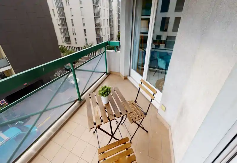 Petit balcon ensoleillé avec un ensemble bistrot en bois et une plante en pot, garde-corps en verre et métal et sol carrelé — parfait pour le café du matin en regardant la rue.