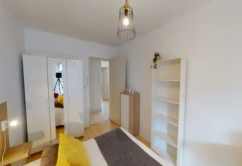 Chambre lumineuse et soignée avec lit double visible, coussins colorés et mobilier blanc simple (armoire et étagères) — idéale pour une annonce locative.
