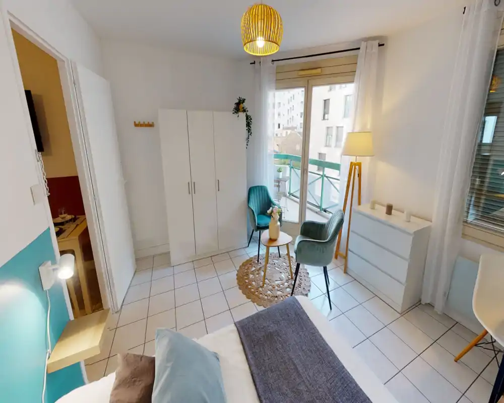 Bright, well-staged bedroom with a comfortable bed in the foreground, a wardrobe, dresser and a small seating area by the balcony letting in natural light.