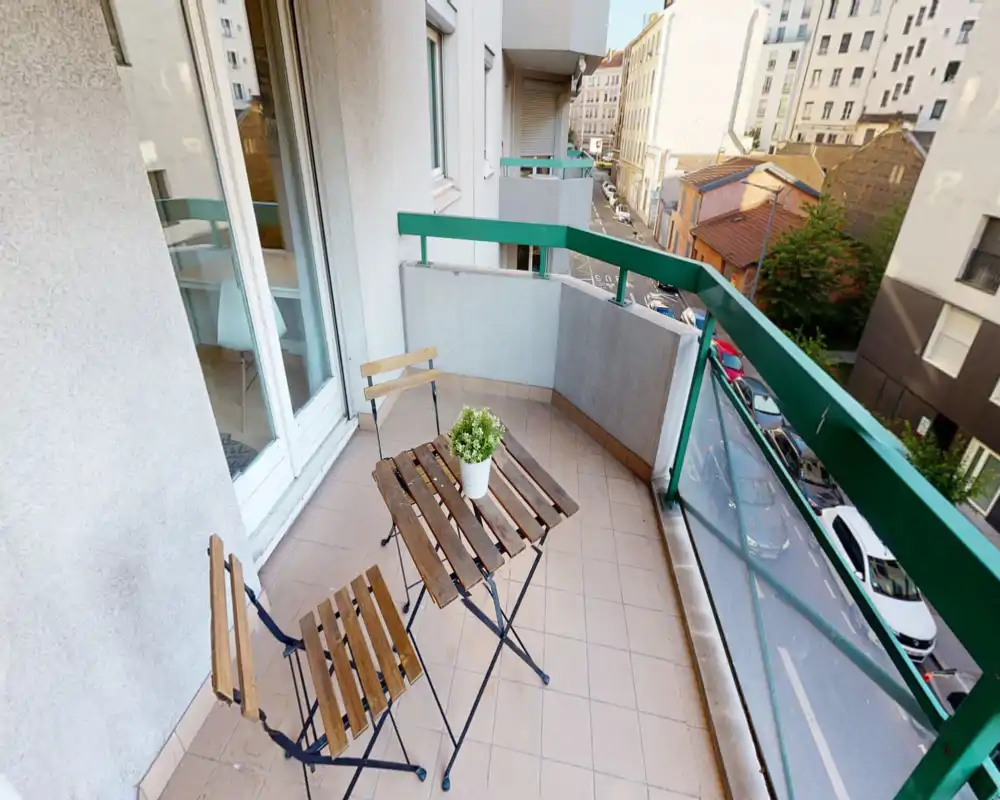 Sunny furnished balcony with a small wooden table, two folding chairs and a potted plant overlooking a quiet city street.