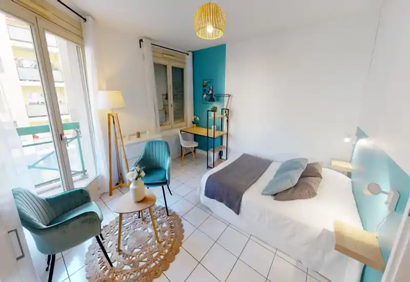 Bright, well-styled bedroom with a double bed, soft pillows and a cozy throw; large window and balcony provide abundant natural light and two comfortable armchairs create a relaxing seating area.