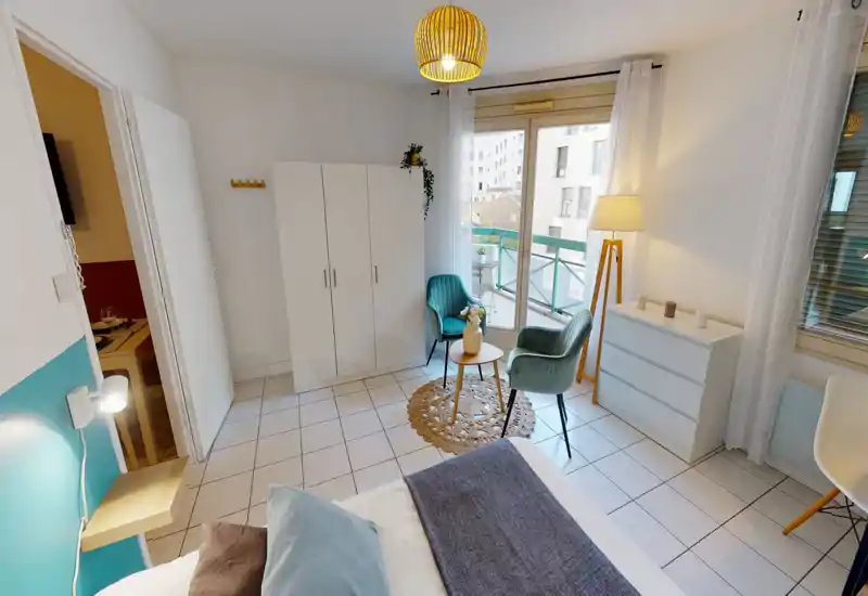 Bright, well-staged bedroom with a comfortable bed in the foreground, a wardrobe, dresser and a small seating area by the balcony letting in natural light.