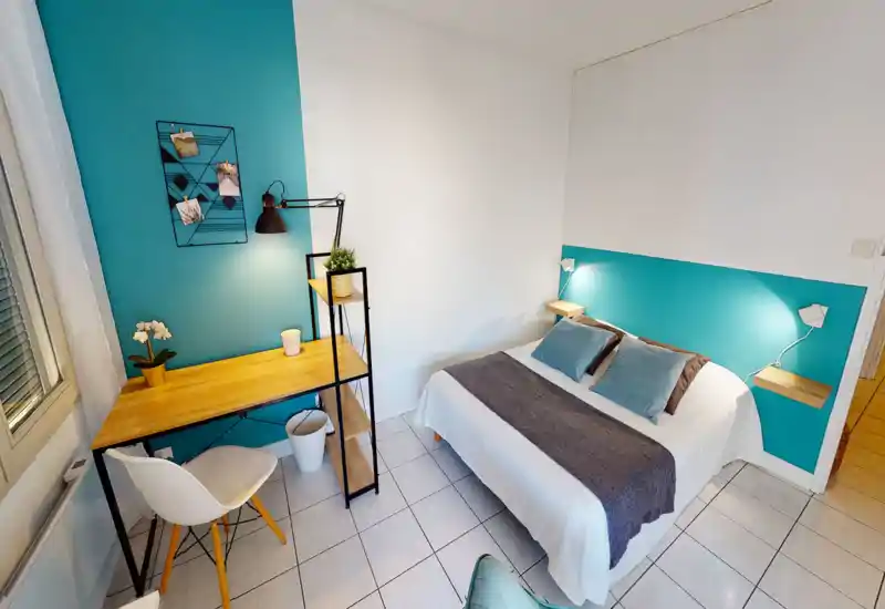 Bright, modern bedroom with a neatly made double bed, coordinated cushions and a bedside shelf lamp. A functional wooden desk with shelving sits by the window, creating a versatile work-and-sleep space.