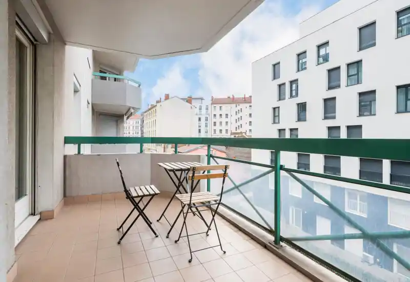 Sunny balcony with a small bistro table and two chairs, tiled floor and glass-and-metal railing overlooking neighboring buildings — ideal for morning coffee.
