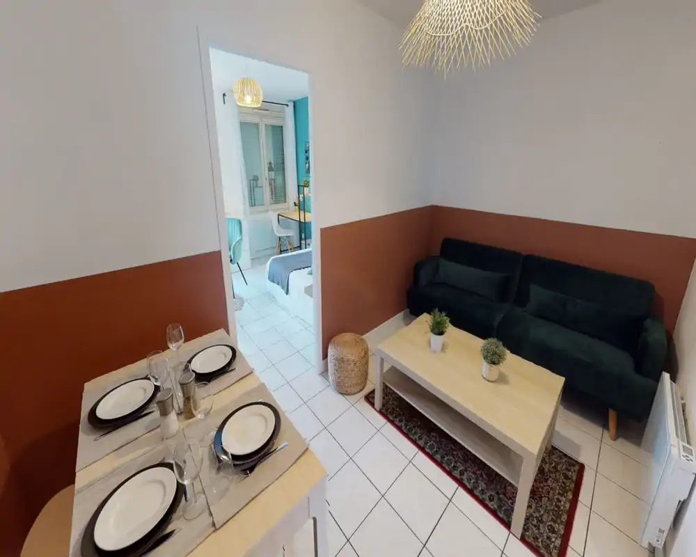 Bright, stylish living area with a dark green sofa, wooden coffee table, decorative plants and a neat dining setup. The space feels welcoming and well-styled with warm wall accents and tiled flooring.