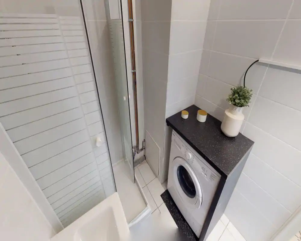 Compact bathroom with a corner shower and an integrated washing machine beneath a black countertop, tiled walls and simple decorative accents.