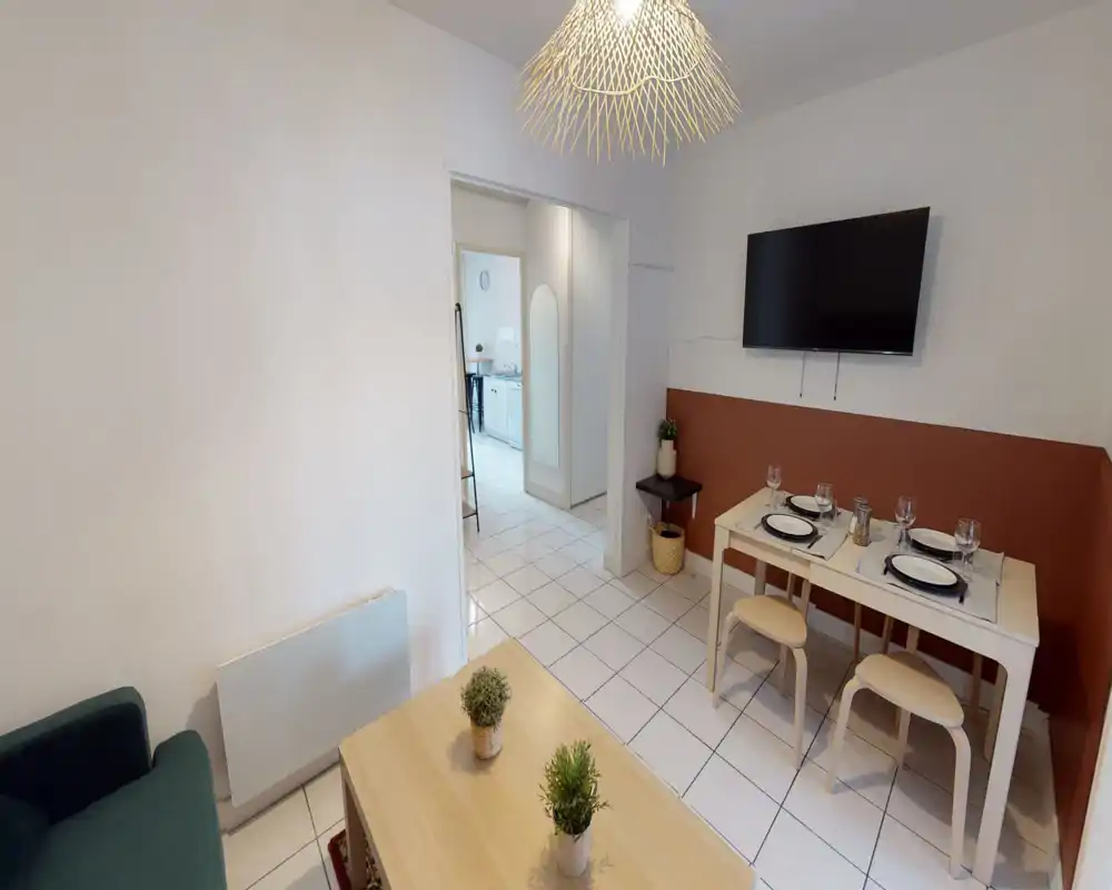 Bright, tidy living area with a small dining table set for four, wall-mounted TV and a low coffee table with plants — comfortable space for relaxing and casual meals.