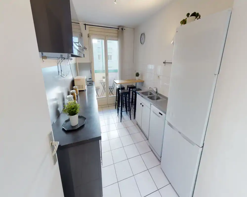 Bright, narrow kitchen with white tiles, full-height fridge, sink and counter space; a small high table and stools face a balcony door that brings natural light.