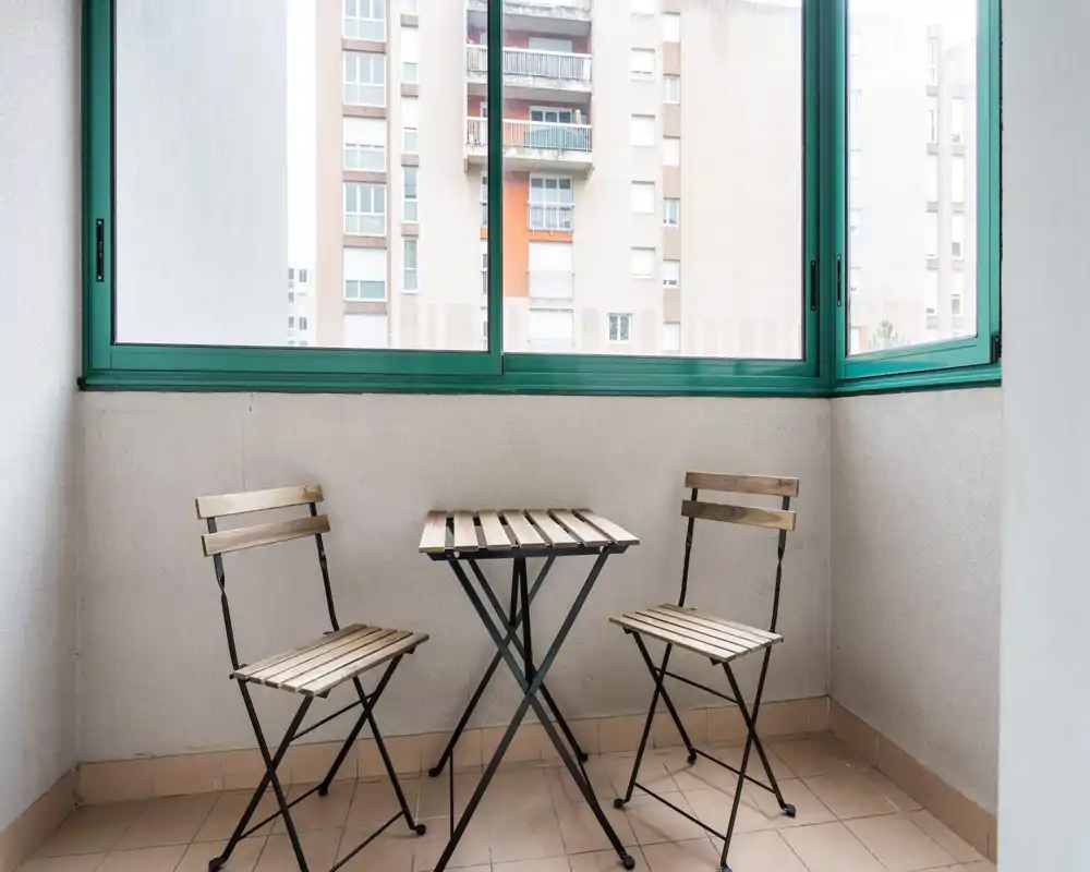 Bright enclosed balcony with a small wooden bistro table and two folding chairs, ideal for morning coffee with an urban view.