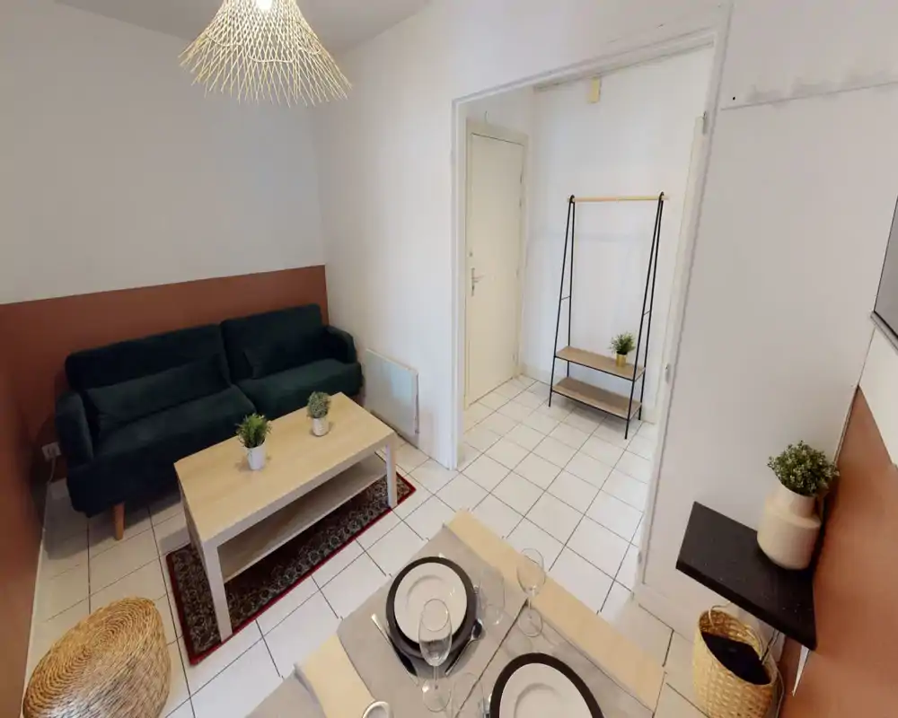 Bright, well-styled living area with a dark green sofa, coffee table, decorative plants and a small dining nook visible in the foreground.