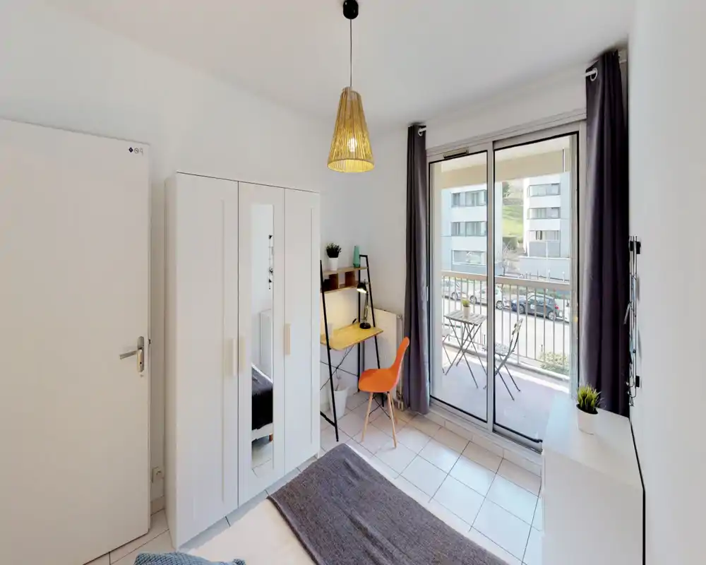 Chambre moderne et lumineuse avec lit visible, armoire blanche avec miroir, petit bureau et porte-fenêtre donnant sur un balcon — agencement soigné et très lumineux.