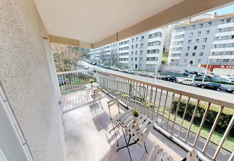 Sunny balcony with small bistro table and chairs, overlooking a street and residential buildings — ideal for morning coffee.