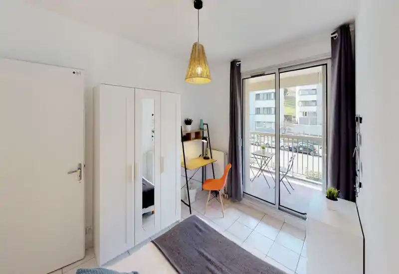 Bright, modern bedroom with a visible bed, white wardrobe with mirror, small desk and sliding doors opening onto a balcony — neat, well-lit and comfortably furnished.